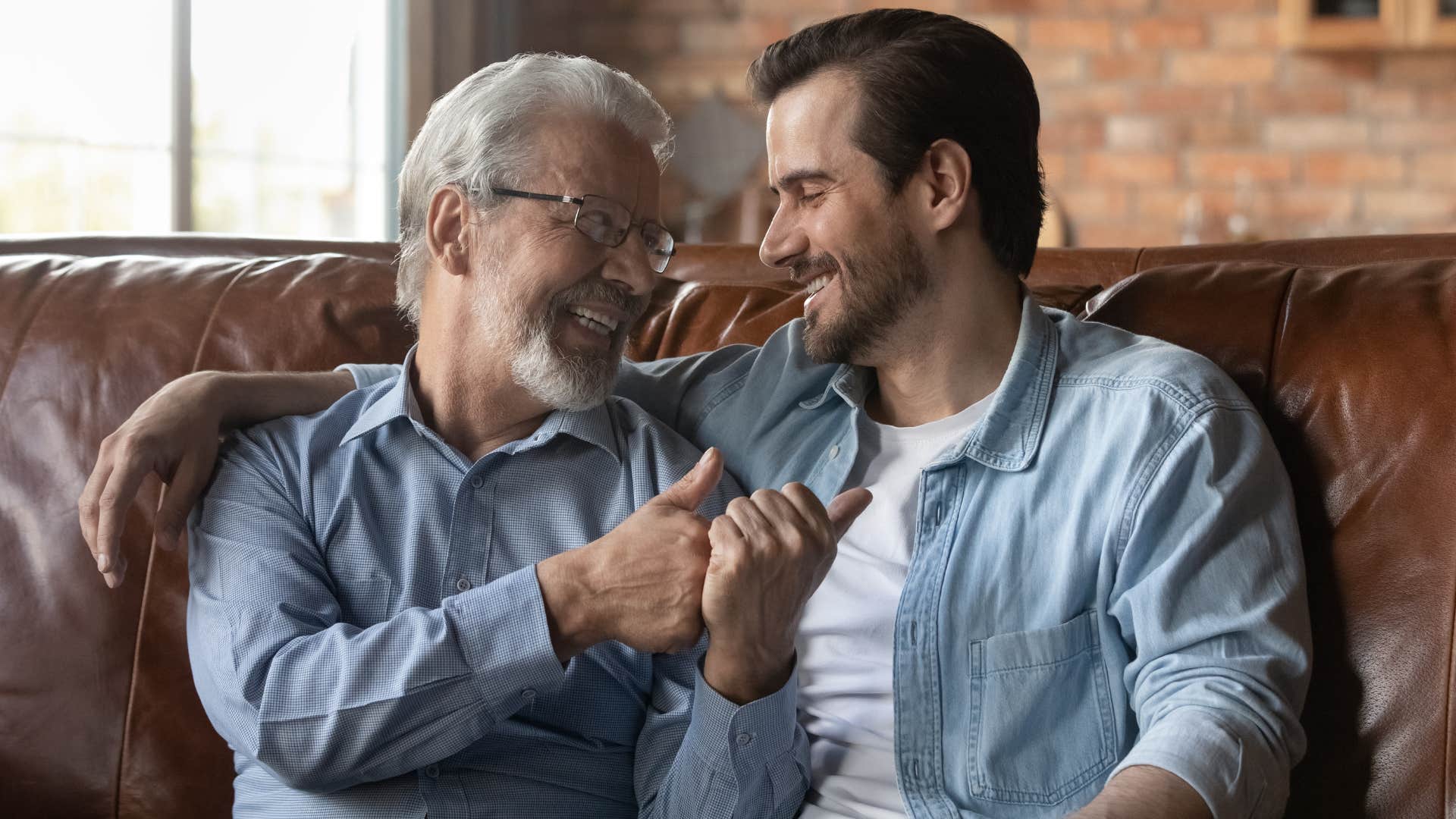 Adult son hugging his dad and smiling on the couch.