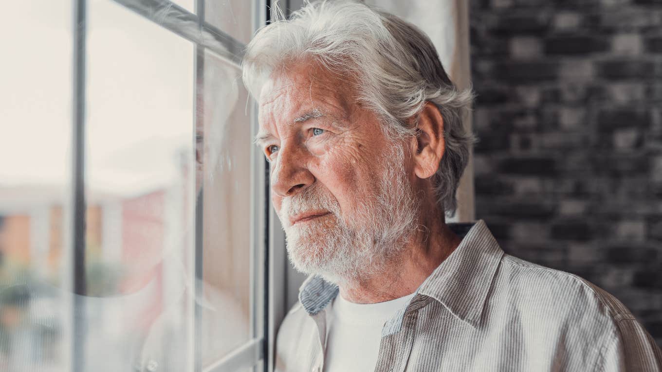 Older man looking sadly out of a window inside.