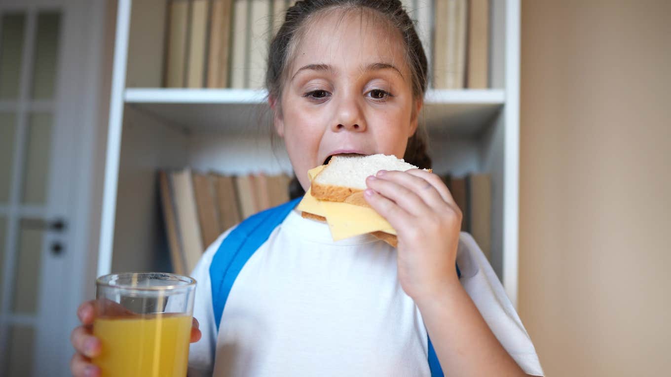 little girl with backpack on eating sandwich and holding glass of juice