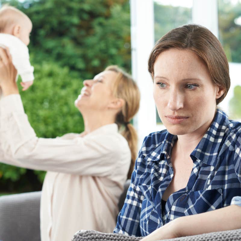 upset woman sitting to the side while friend plays with baby