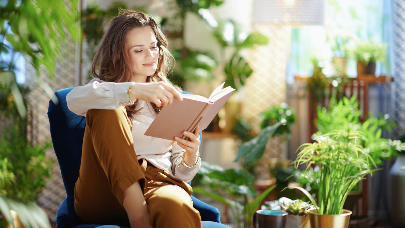 relaxed trendy middle aged woman with long wavy hair with book in green pants and grey blouse in the modern living room in sunny day.