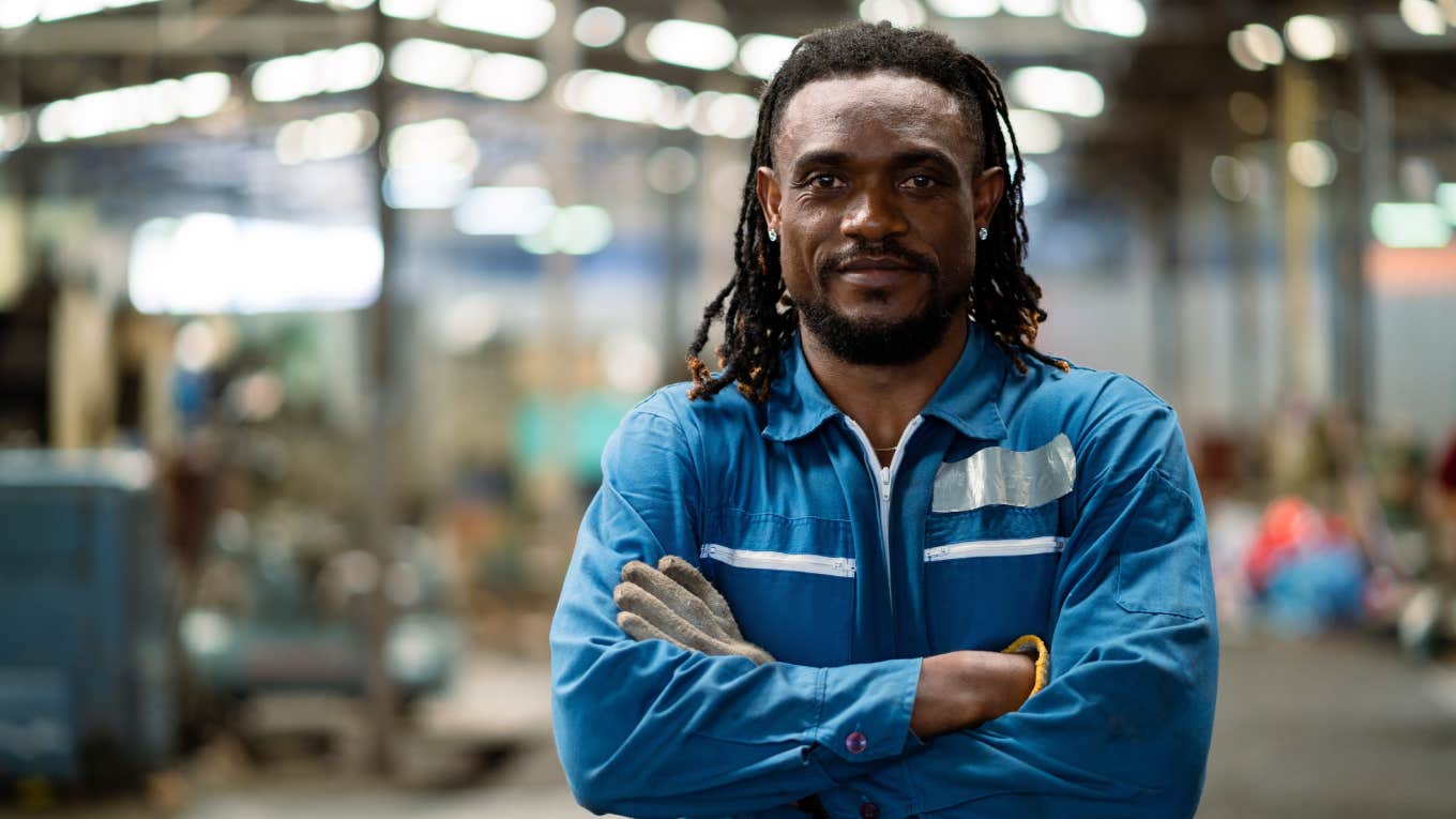 man in a blue uniform stands in a factory with his arms crossed.