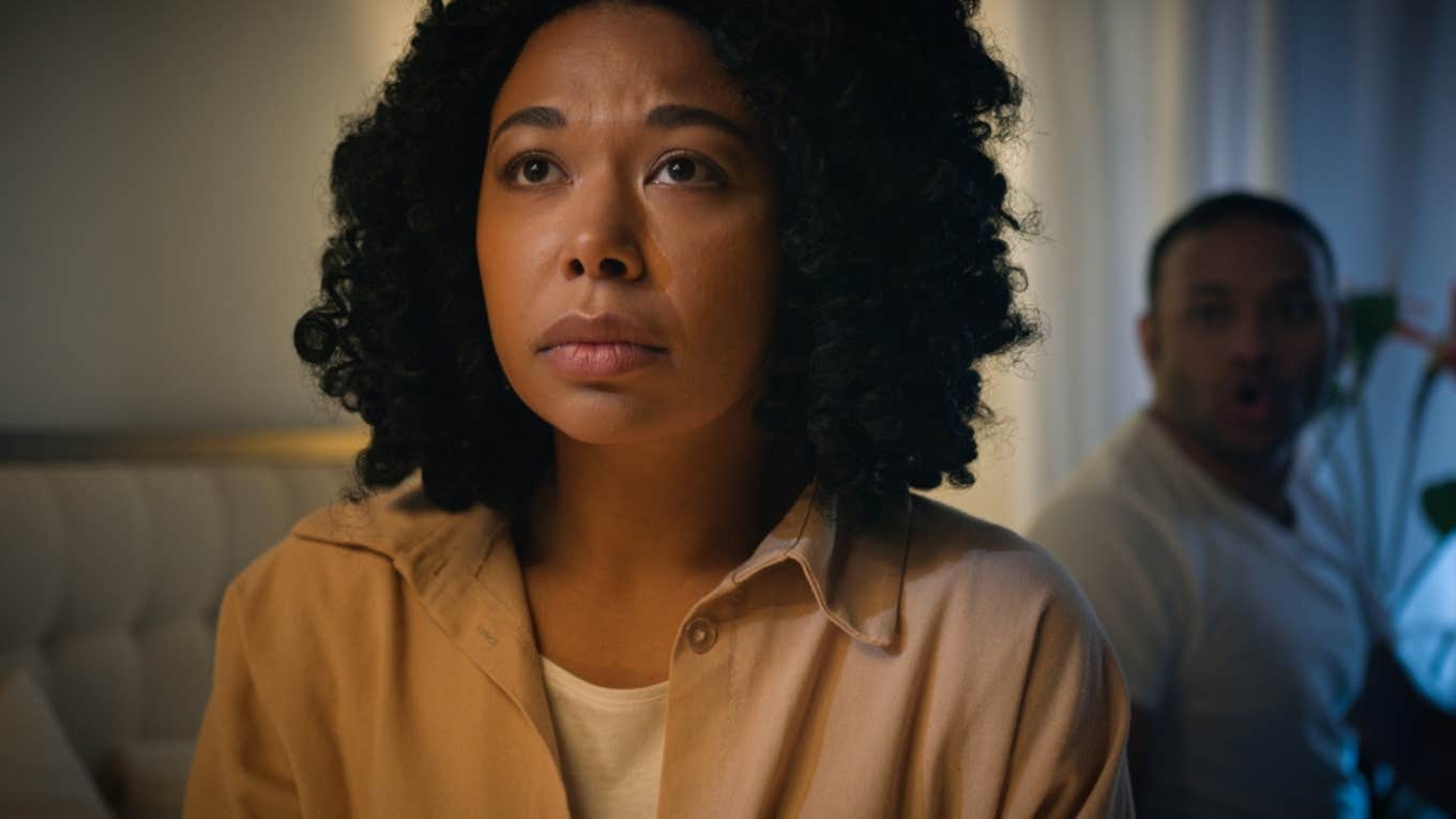 Woman looking solemn next to an angry man behind her.