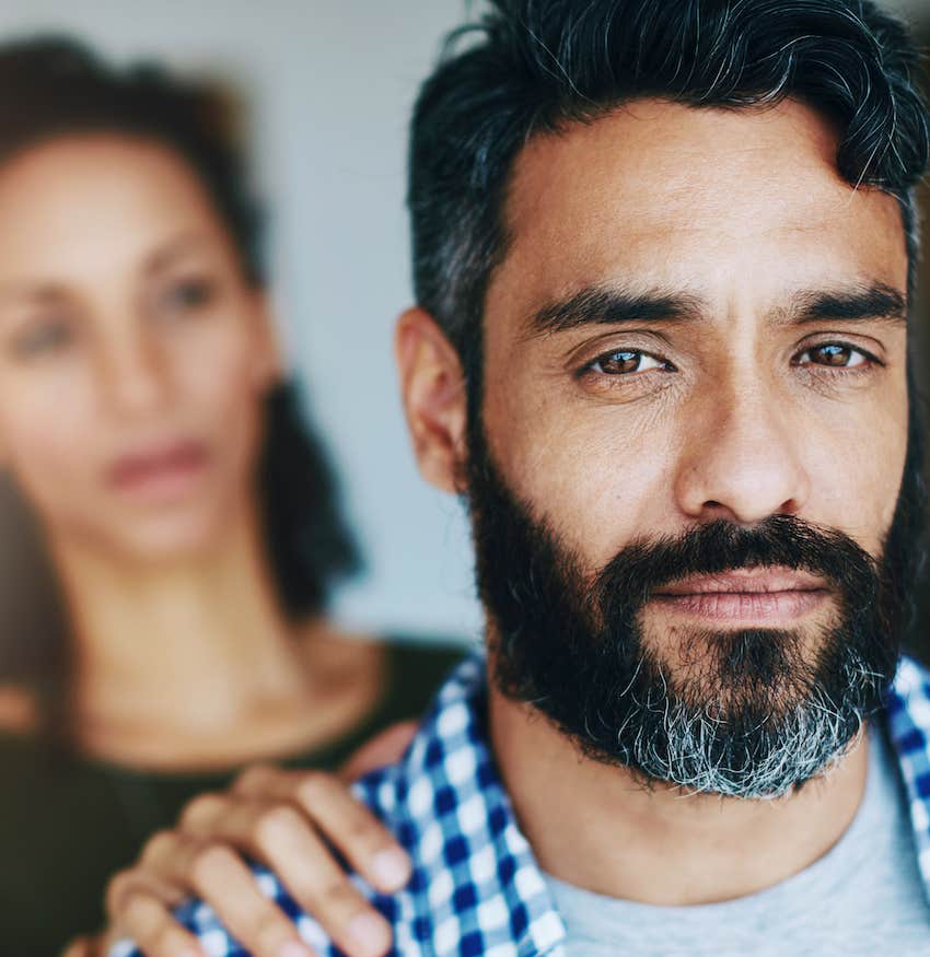 Upset man looks at camera, from behind, a woman touches his shoulder 