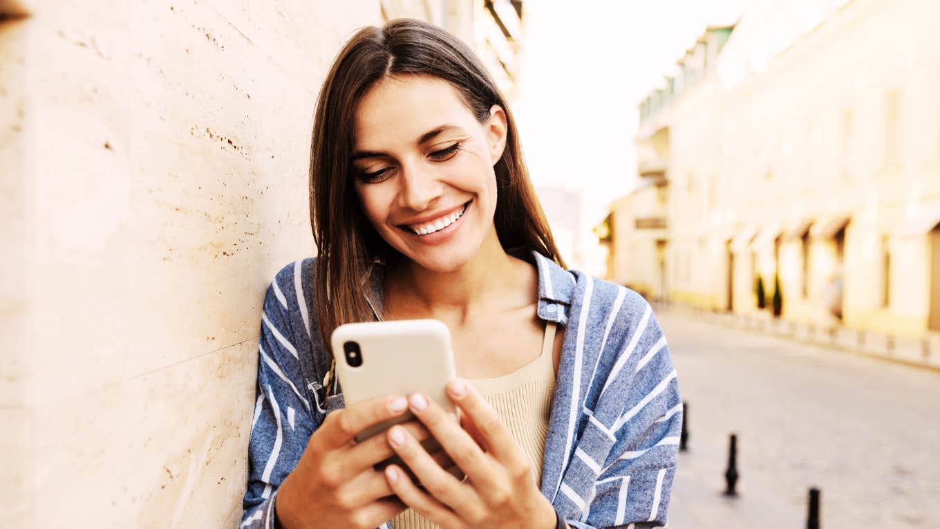 Woman leaning on wall smiling big, texting.