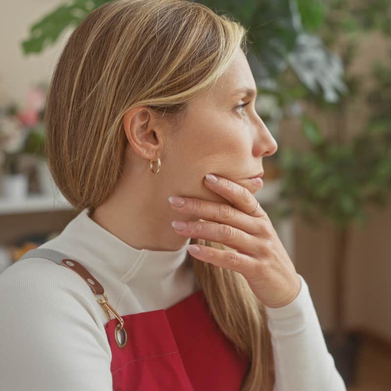 A contemplative young woman stands indoors surrounded by vibrant foliage in a flower shop.