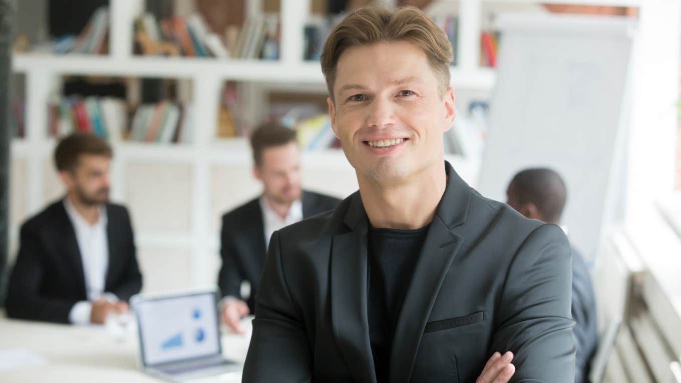 Smiling confident executive in suit standing with arms crossed looking at camera with business partners team at background