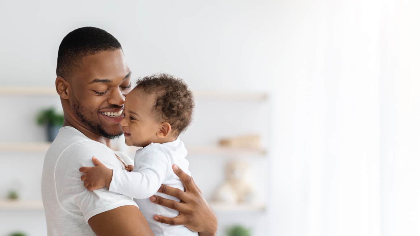 Portrait Of Happy African American Father With Infant Baby In Arms Standing Near Window At Home