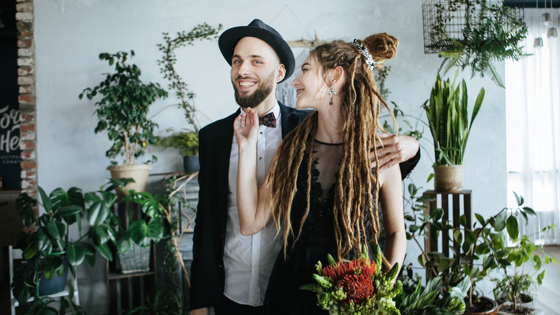 a stylish woman in a black dress and with dreadlocks and a man in a black hat hugging against a background of exotic plants