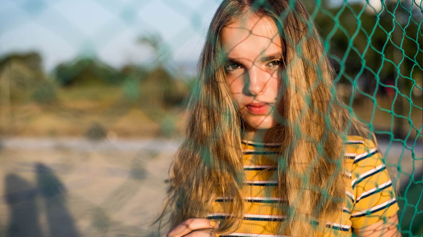 young woman with long hair and striped shirt looking through a chain link fence