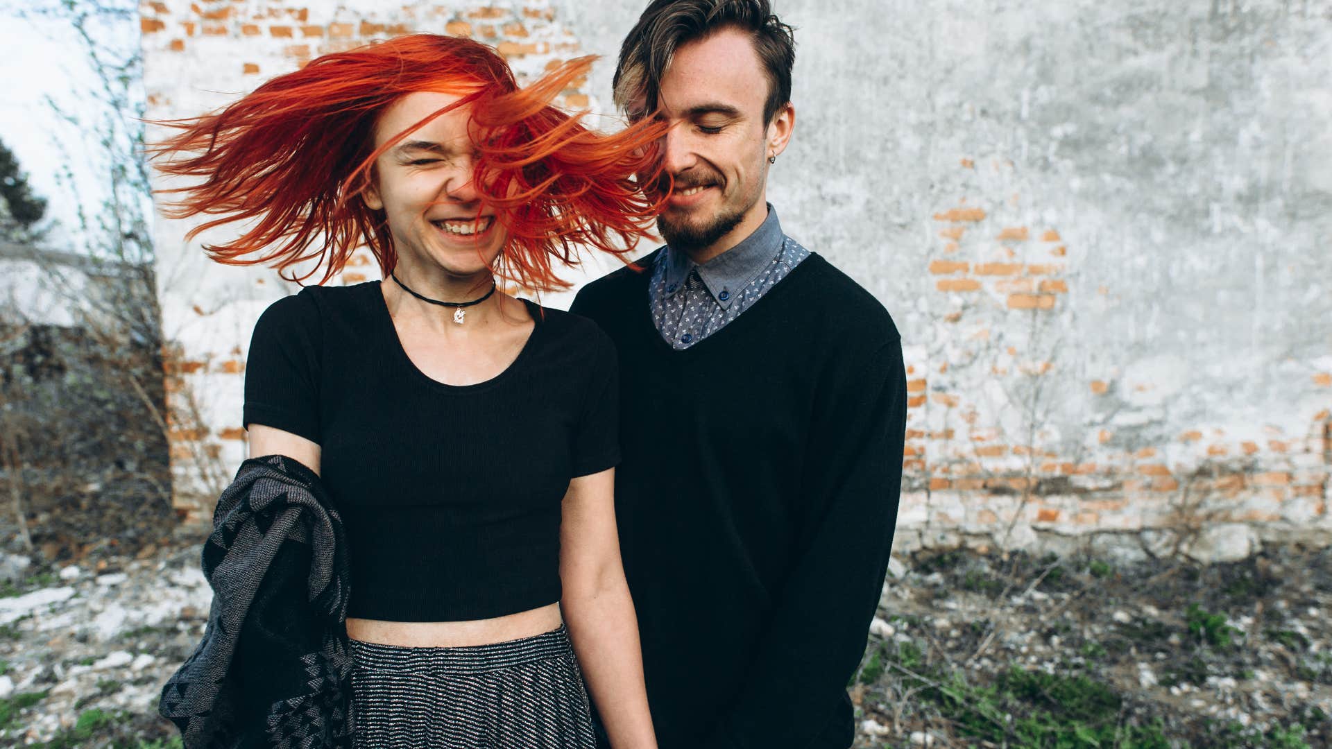 woman with bright red hair laughs with her partner by a white wall