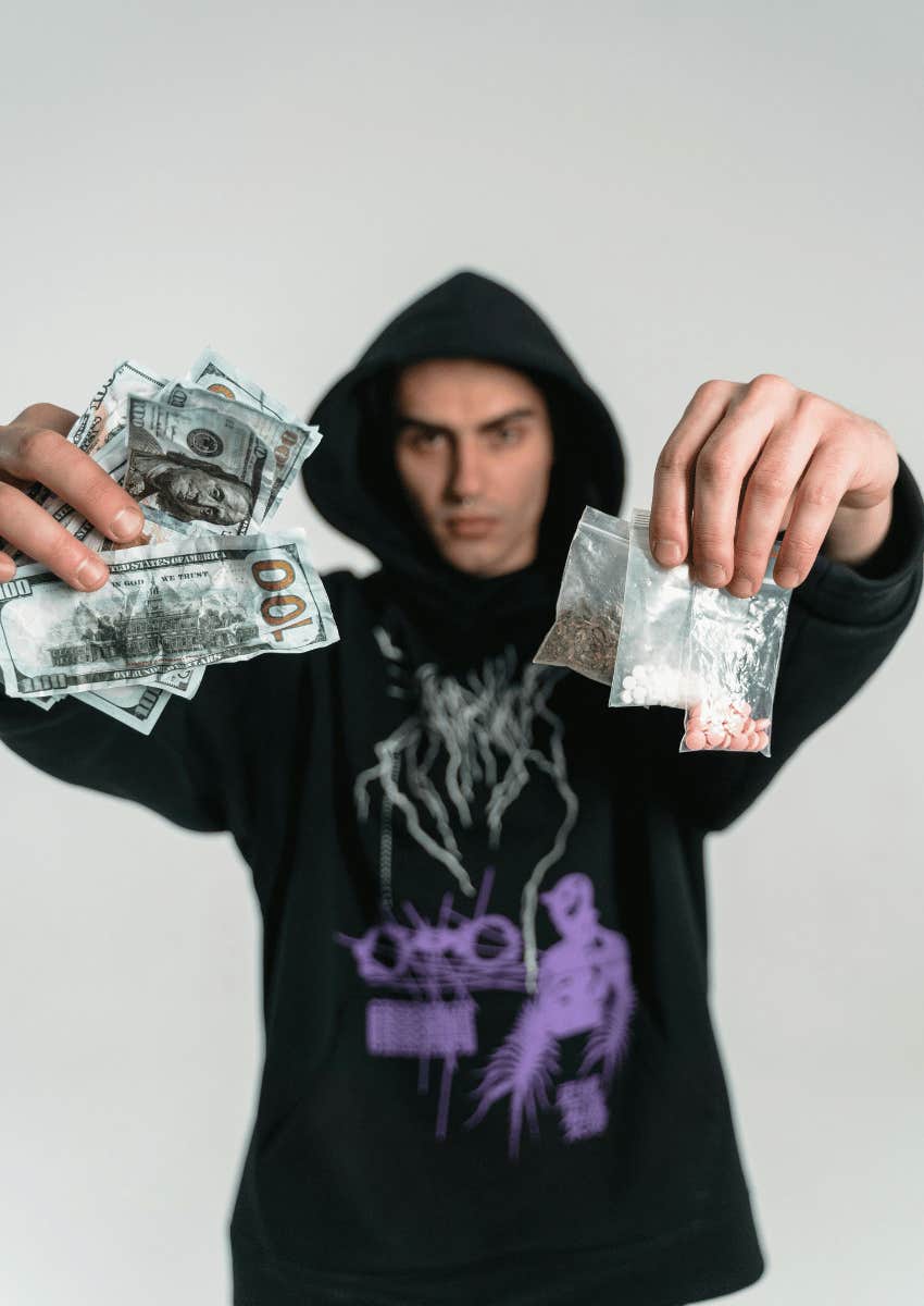 man in a hood holding money and small bags of pills