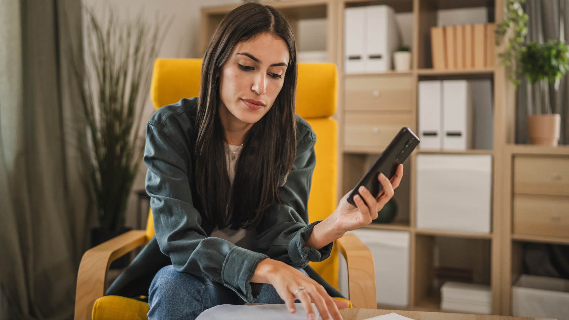 Young woman holding her phone and looking at papers on a table.