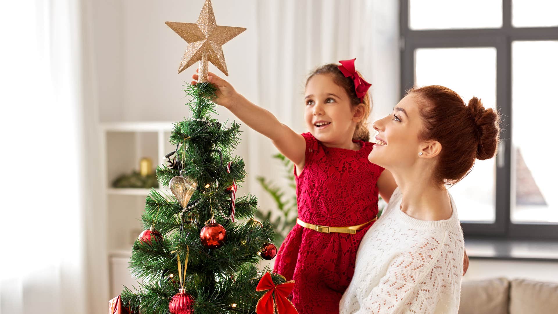 Woman holding her young daughter while putting up a Christmas tree.