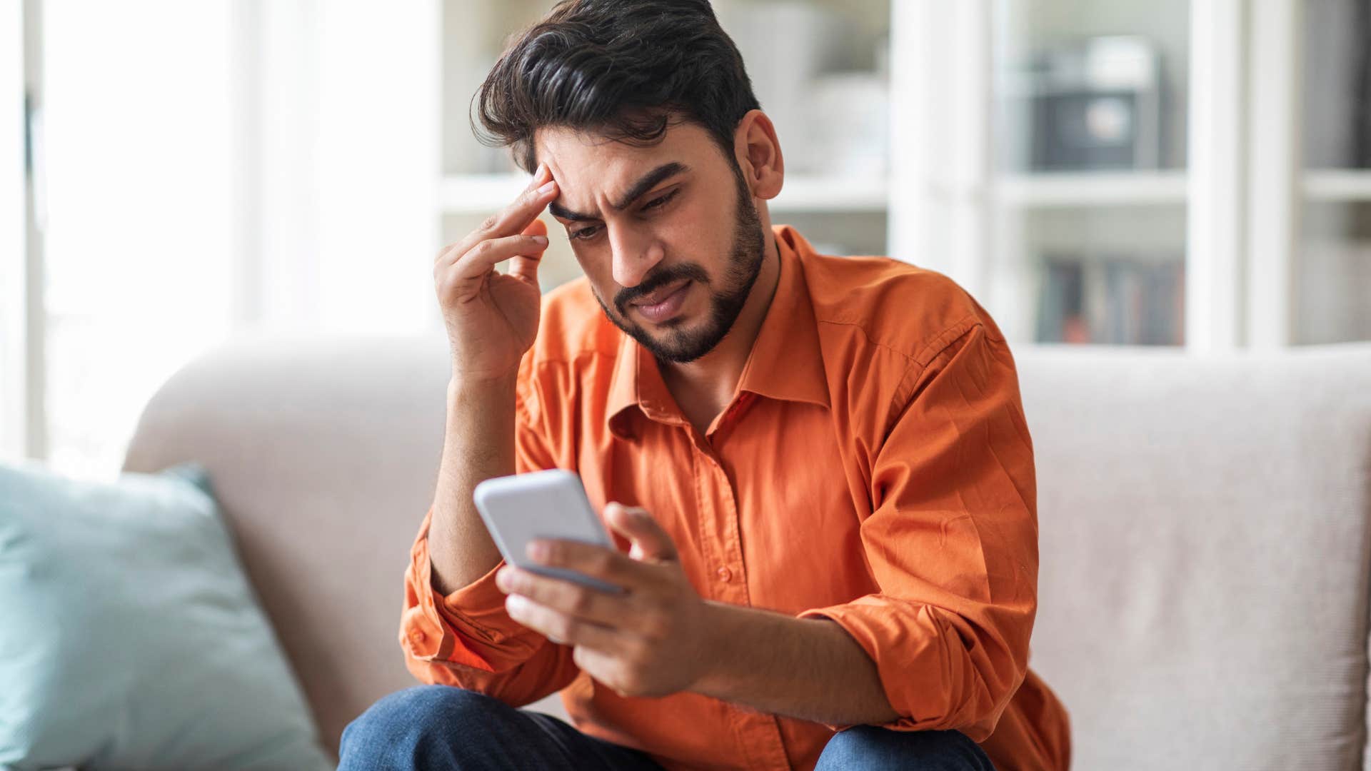 Man wearing an orange shirt looking confused at his phone.