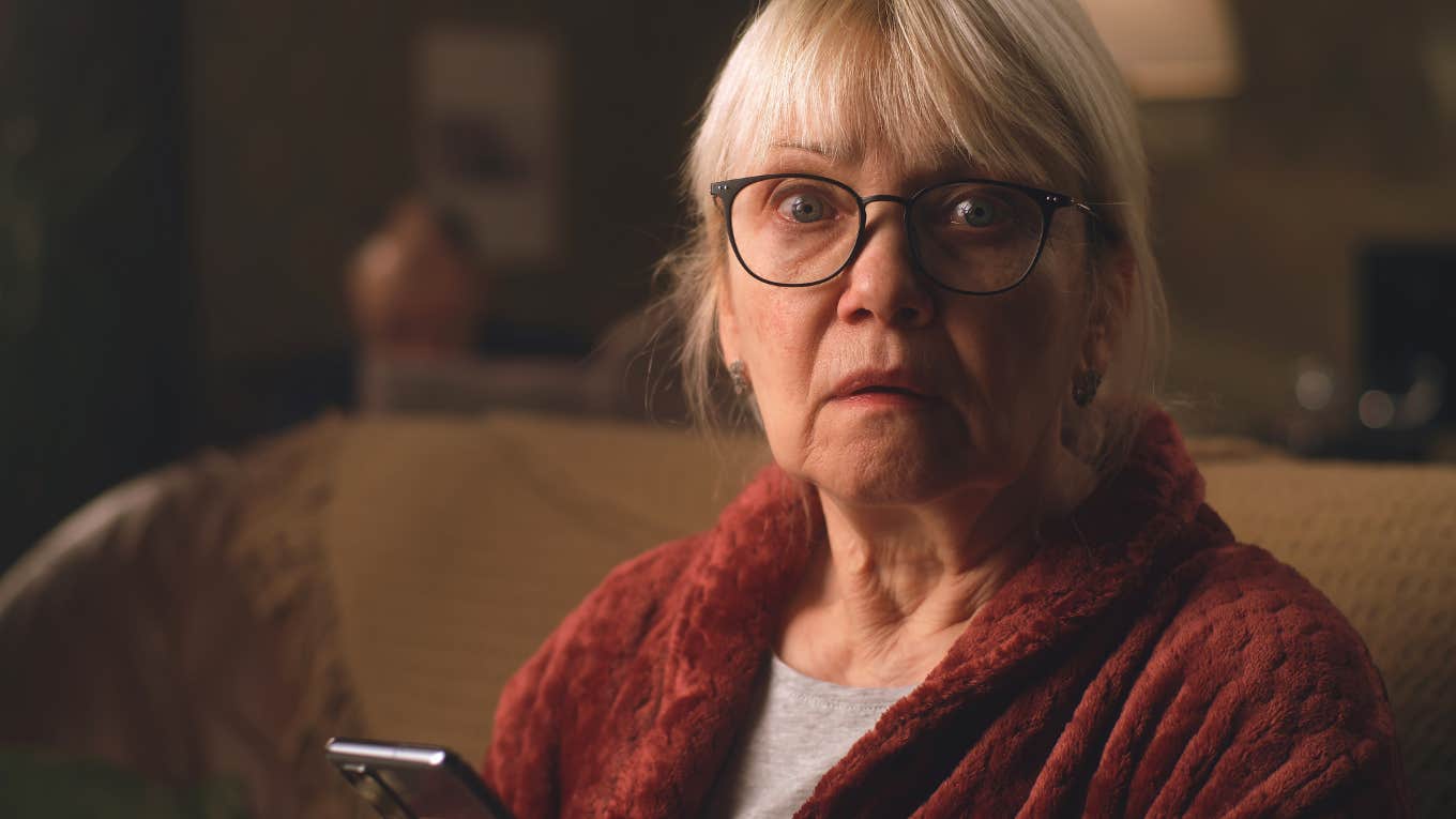 Shocked elderly female in glasses looking around after reading on mobile phone while sitting on sofa in living room at home