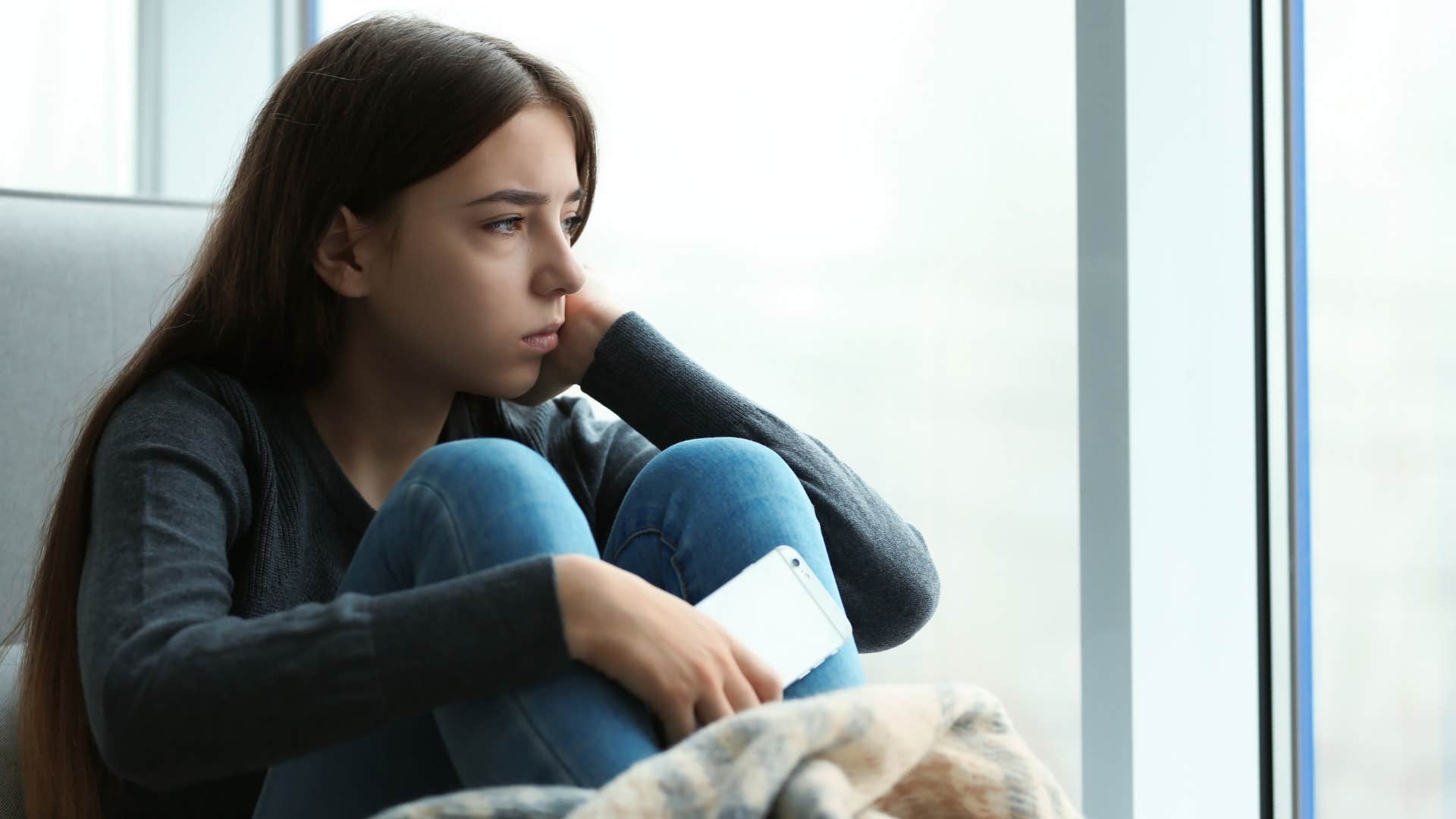 Teenage woman looking sad staring out a window.