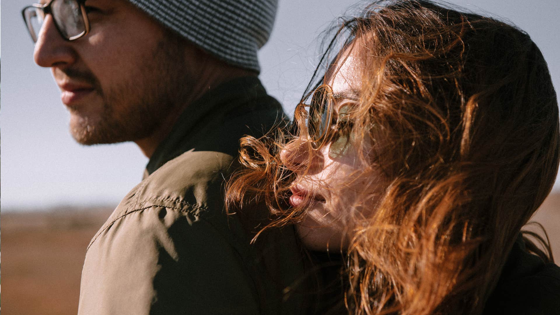 brown haired woman with sunglasses rests her chin on the shoulder of her partner with a beanie on his head