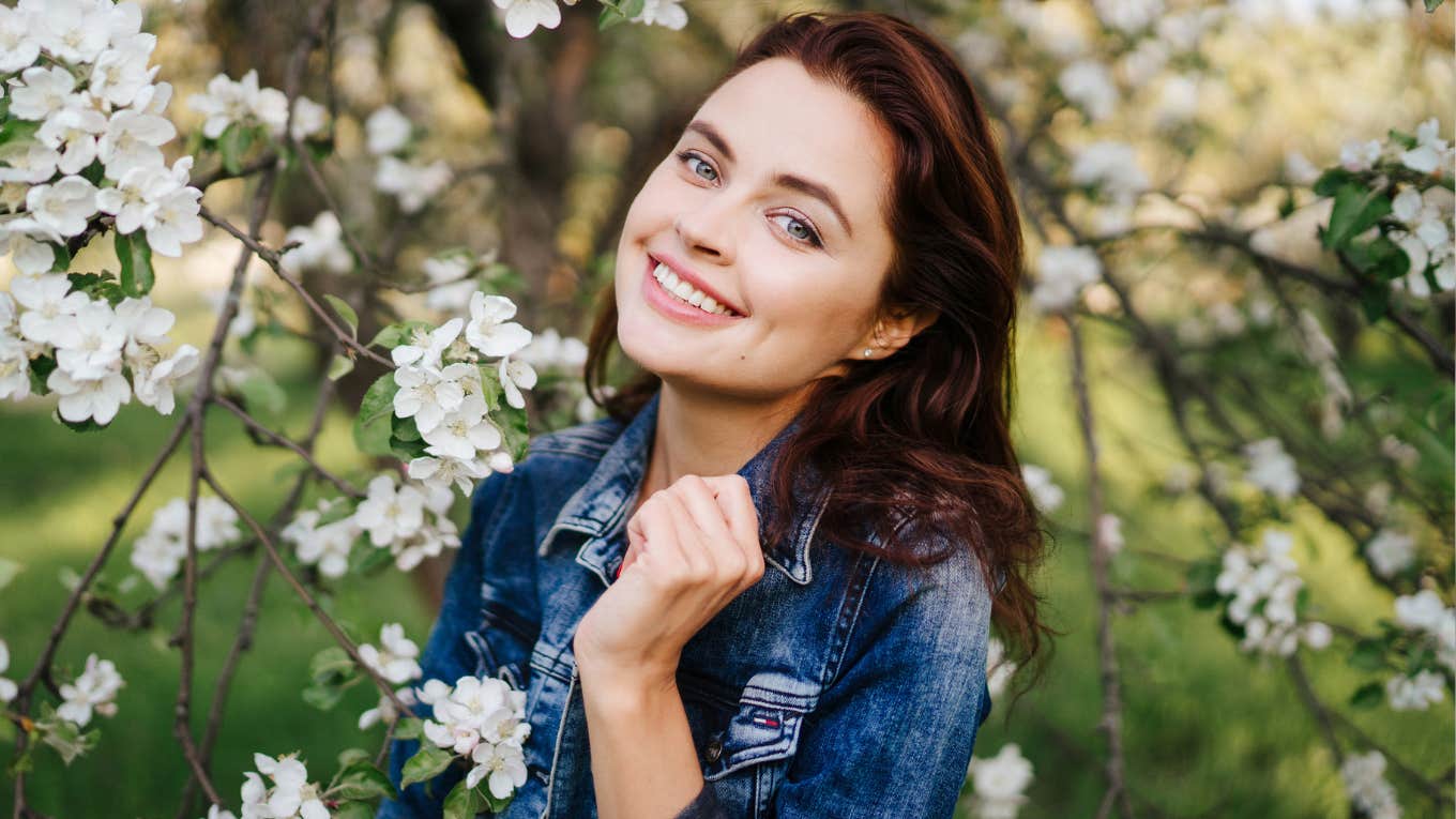 Young smiling woman in jeans jacket standing near blooming spring tree. 