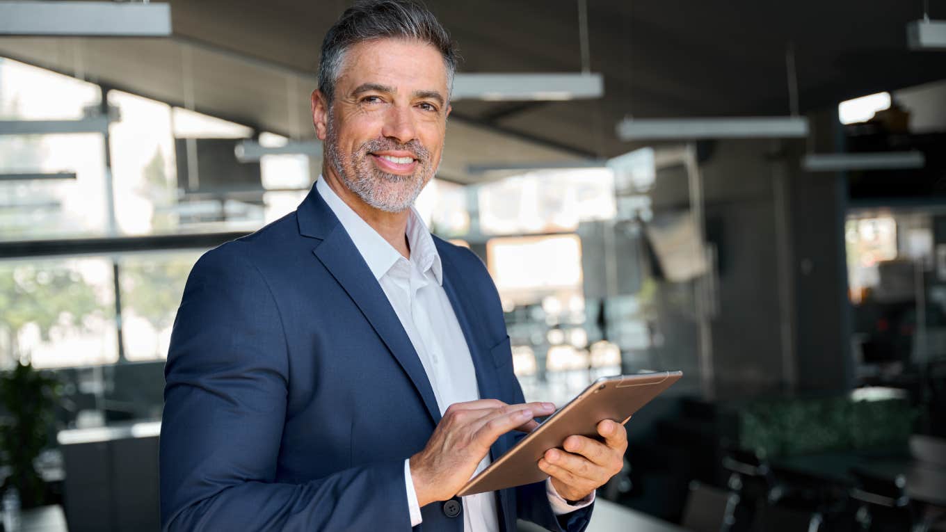 Happy middle aged ceo wearing suit standing in office using digital tablet