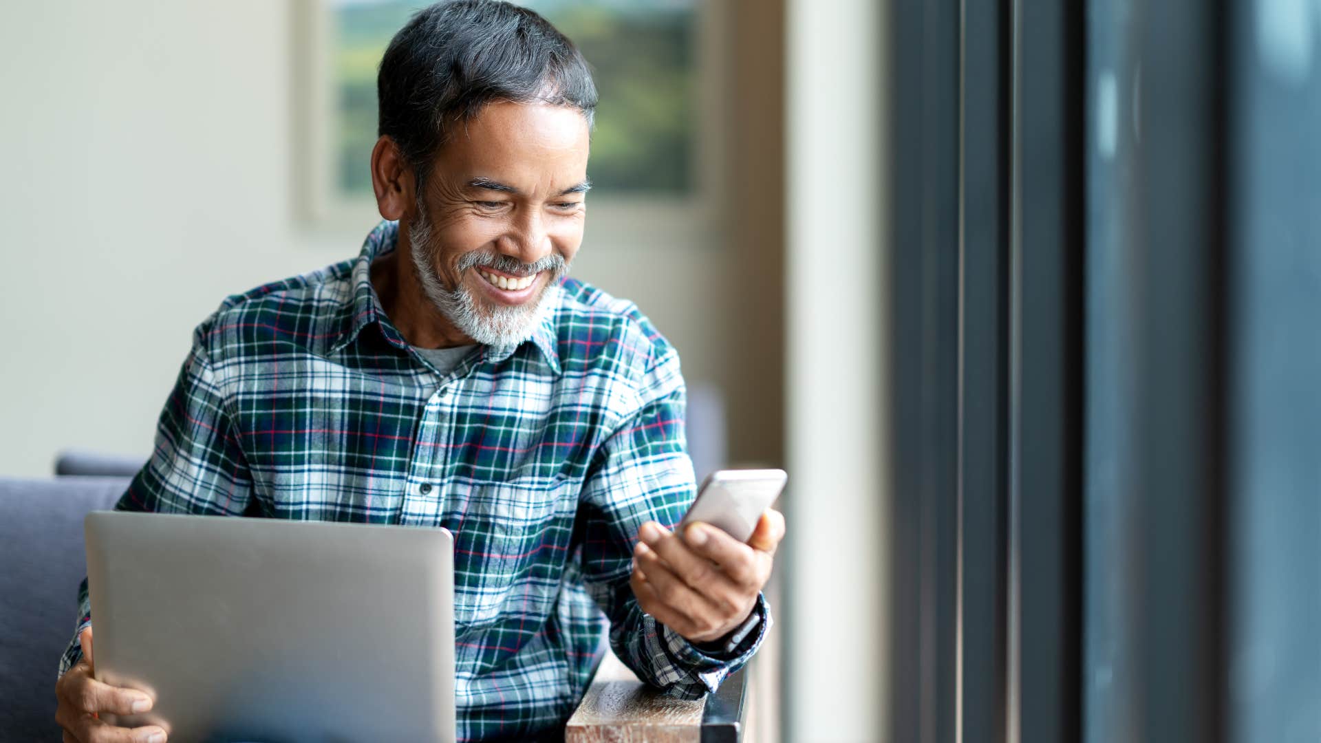 Adult man smiling and looking at his phone and laptop