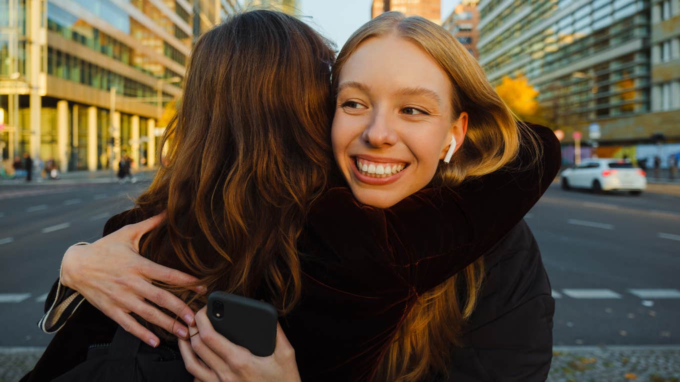 Smiling young woman in earphones hugging her friend when they meet at city street