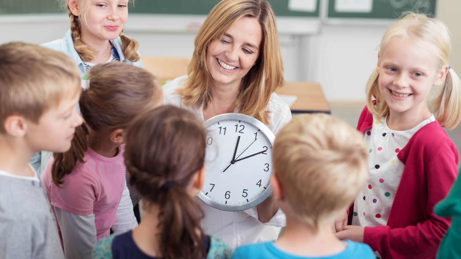 Teacher holding an analog clock surrounding by young kids.