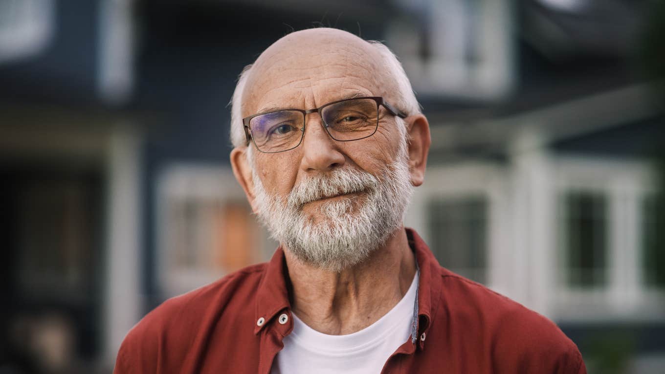 older man smirking standing outside of his house