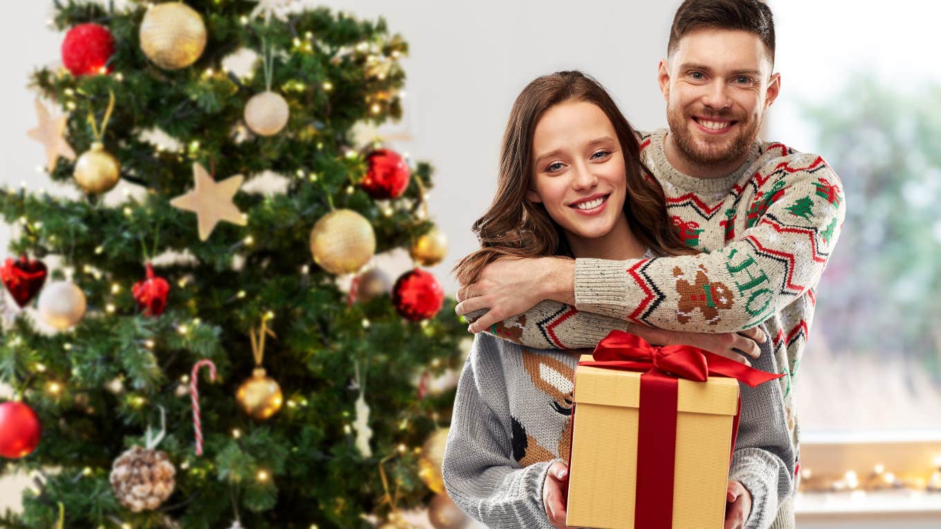 couple holding a gift standing next to a Christmas tree