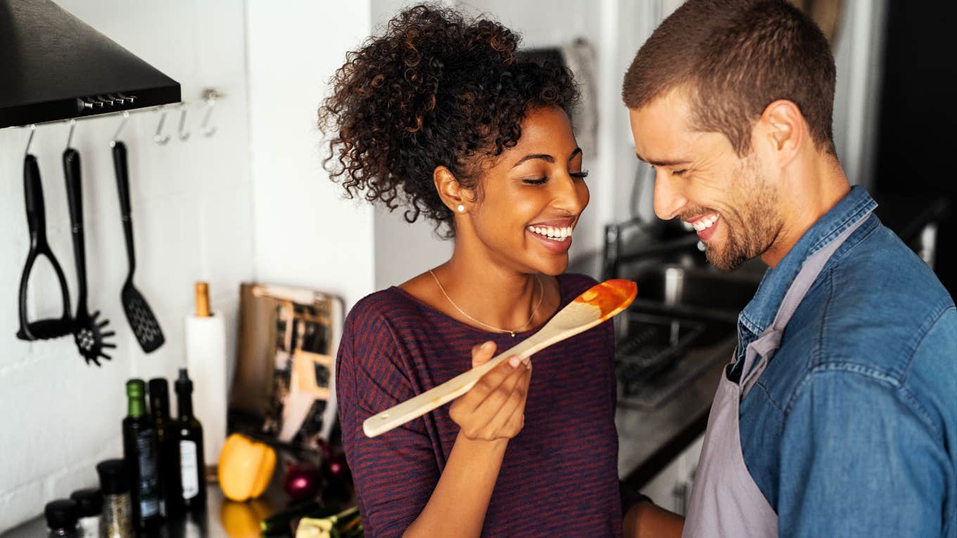 Couple cooking Italian food in kitchen, laughing taste testing red sauce. 