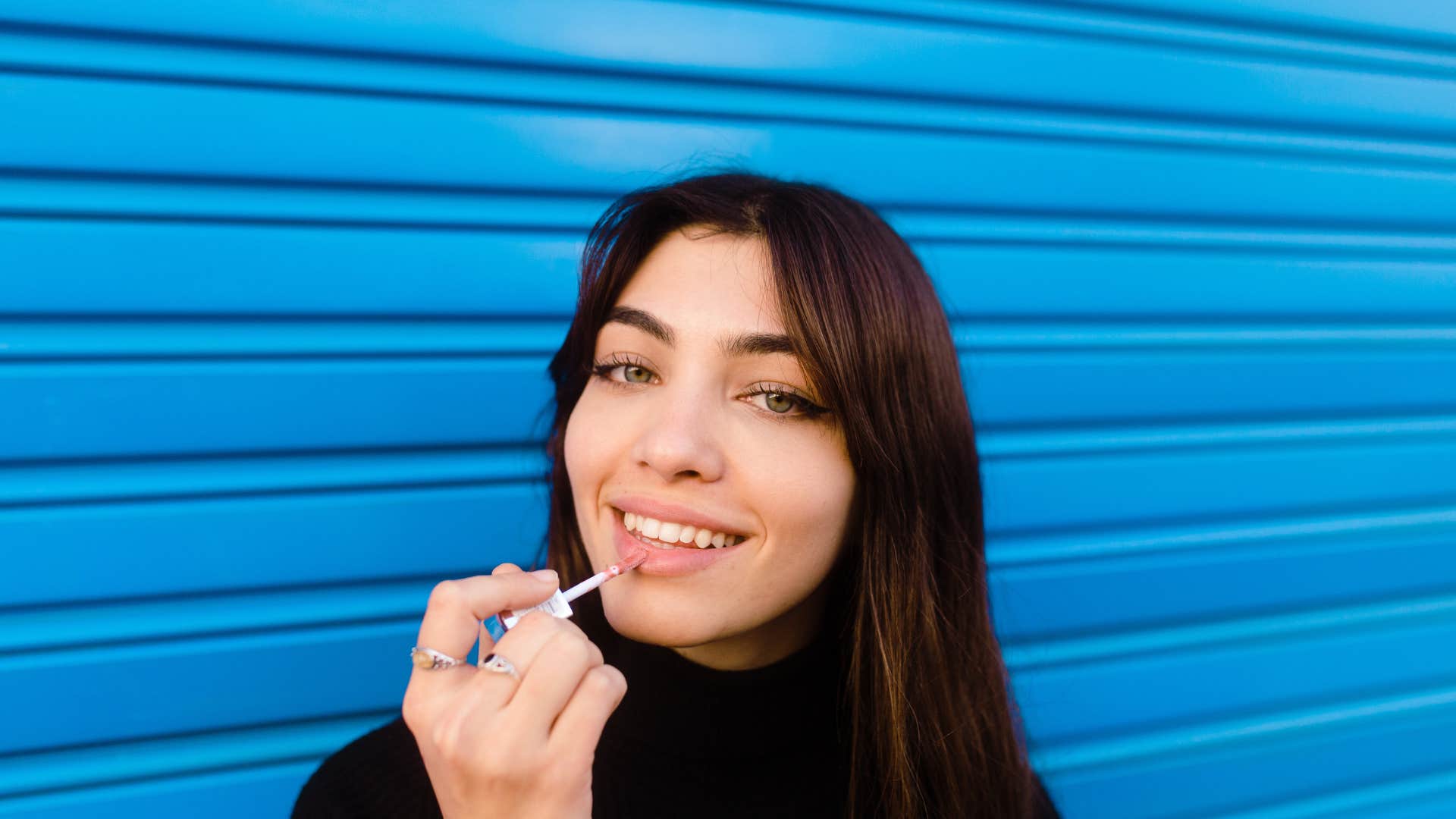 brunette woman smiling and applying lip plumping gloss in a blue urban background 