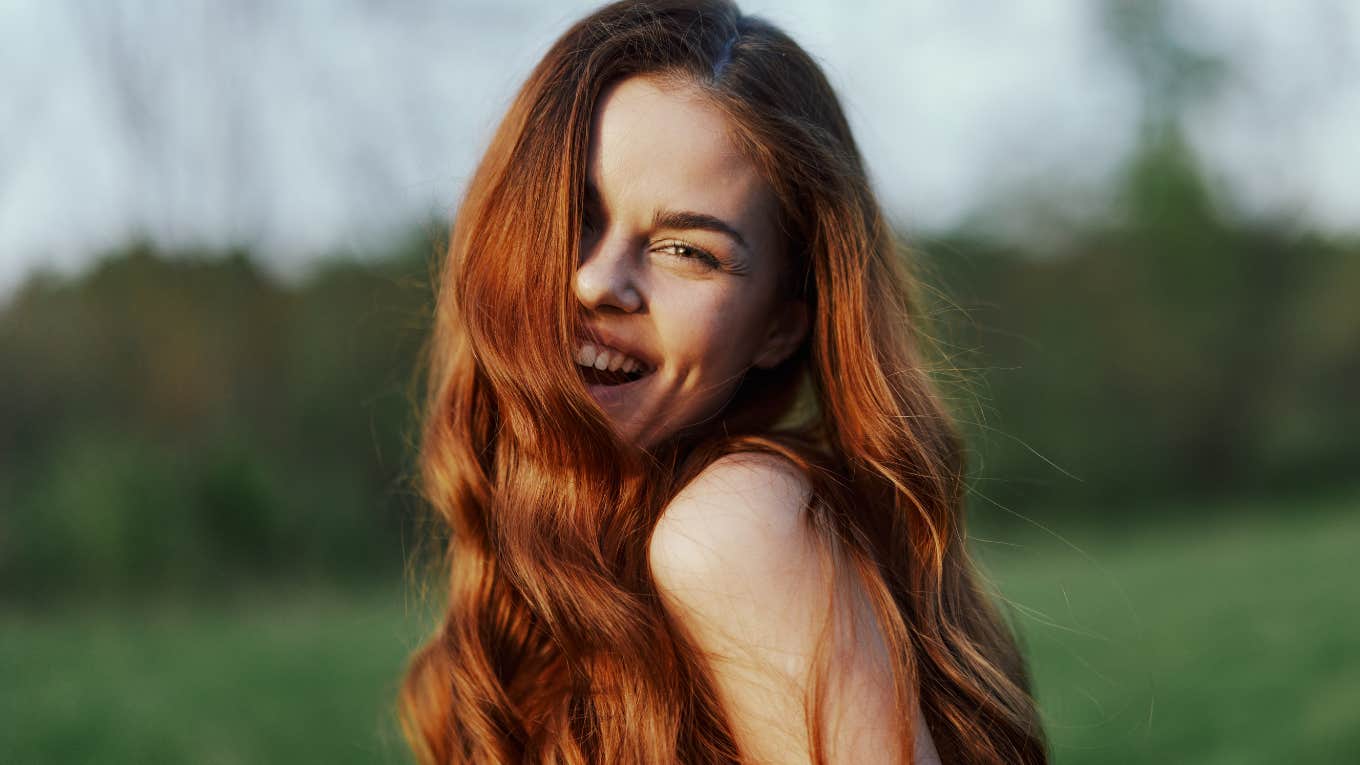 A young woman smiles and looks into the camera with her long, red, wavy, shiny hair in a park with green grass