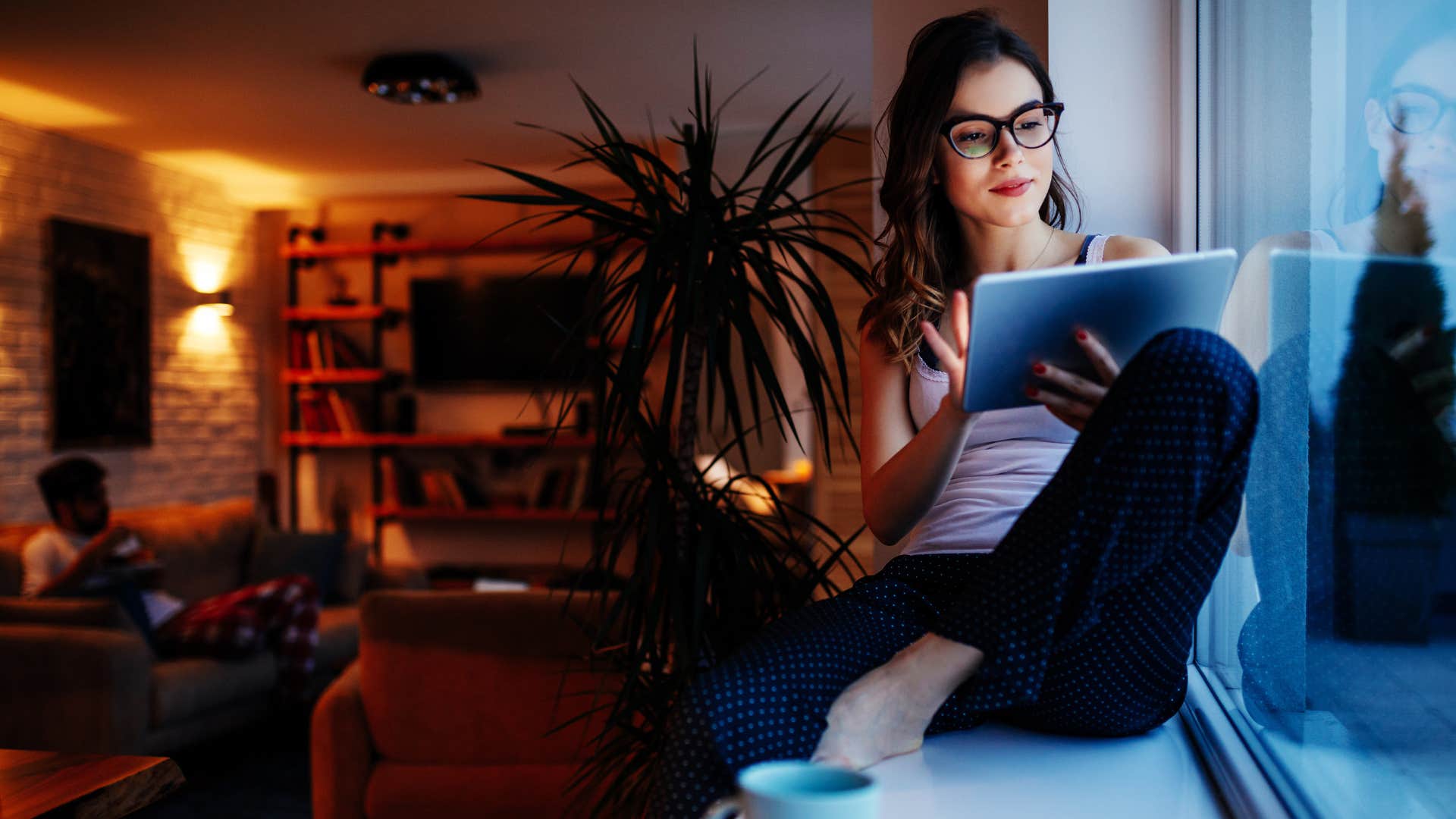 Photo of a young woman sitting by the window and using digital tablet at home.