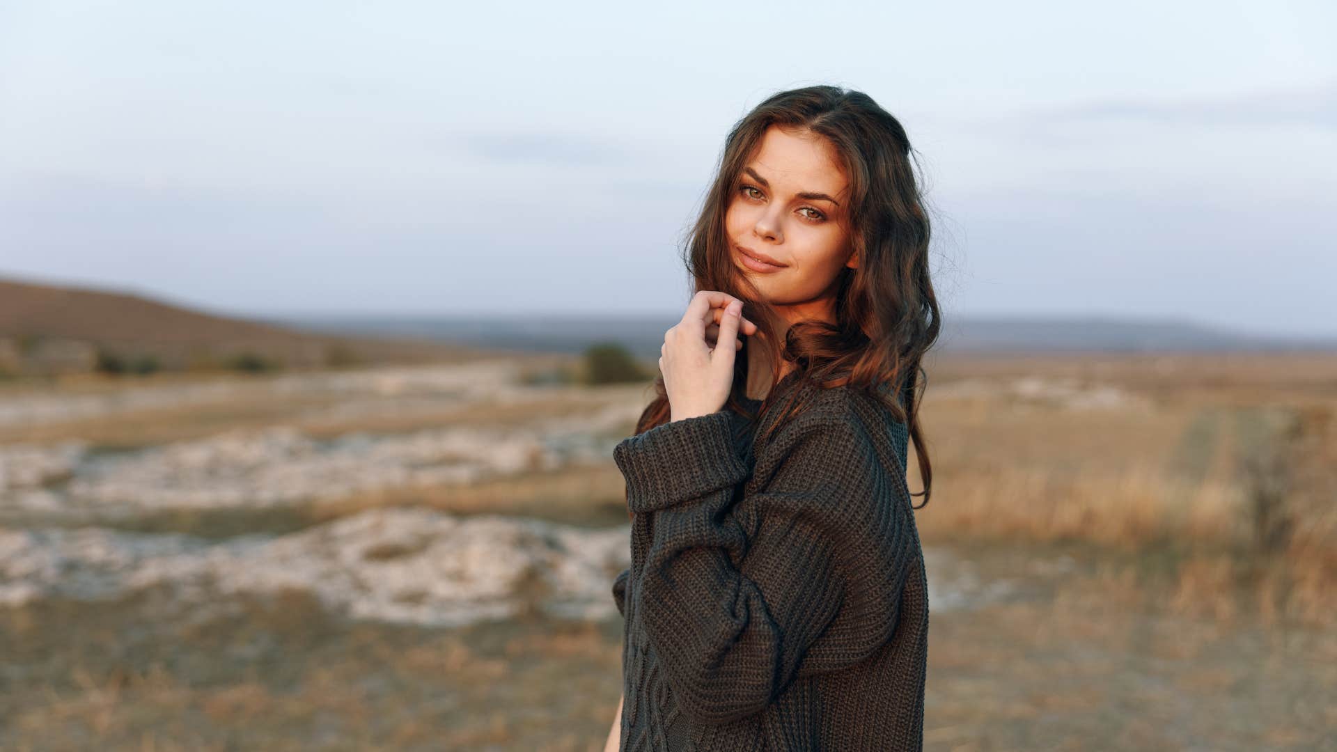 Serene young woman in cozy sweater standing under the golden sunset in vast field