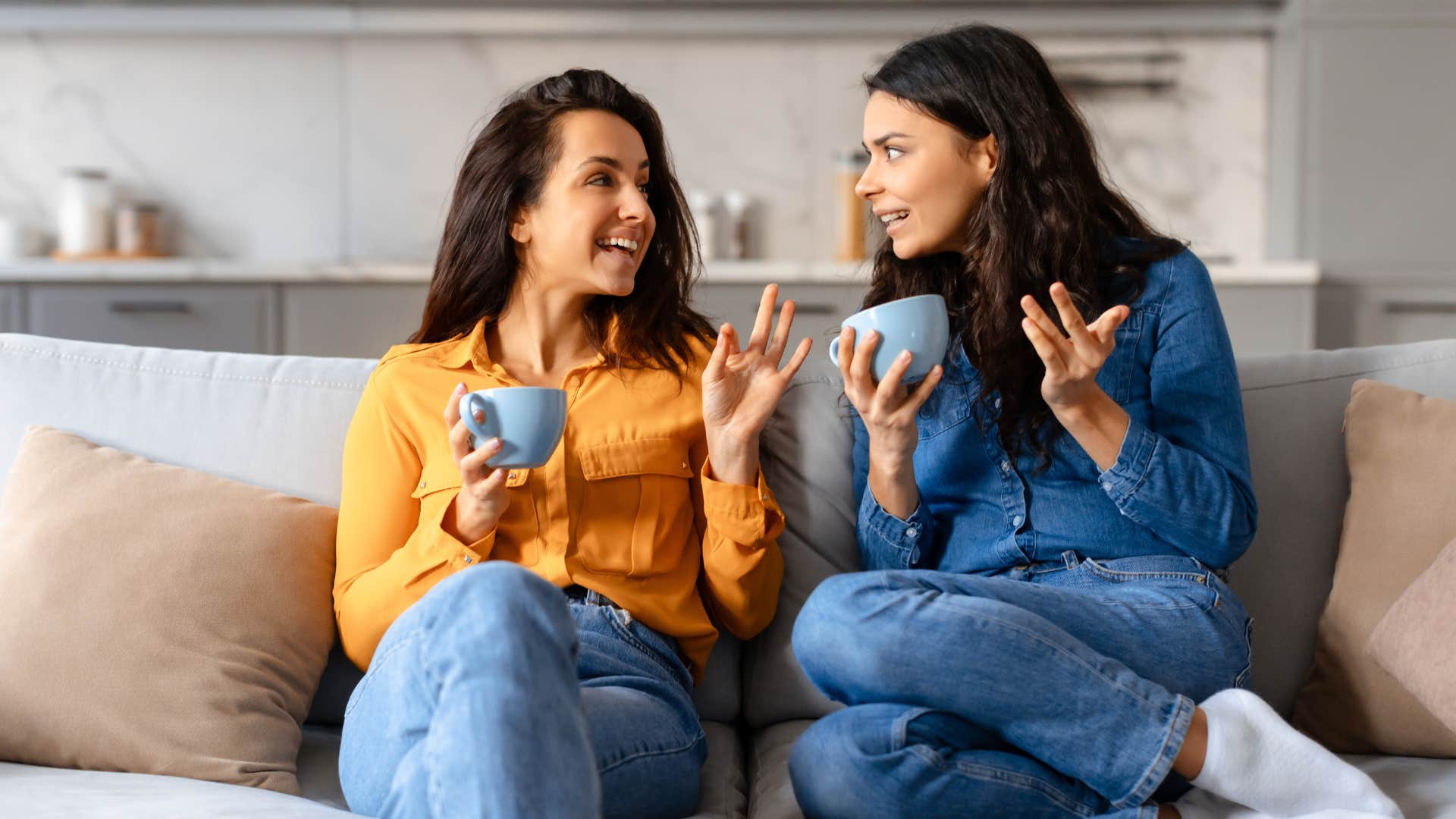 Two women talking with coffee mugs on the couch.