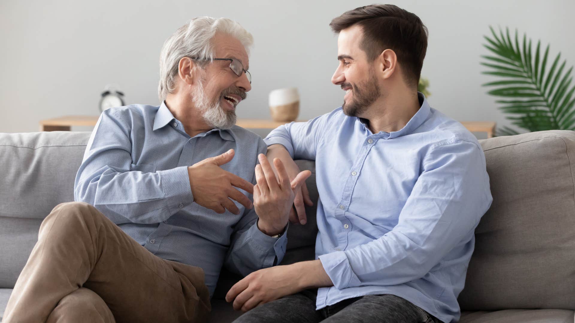 Adult son and father smiling and talking on the couch.