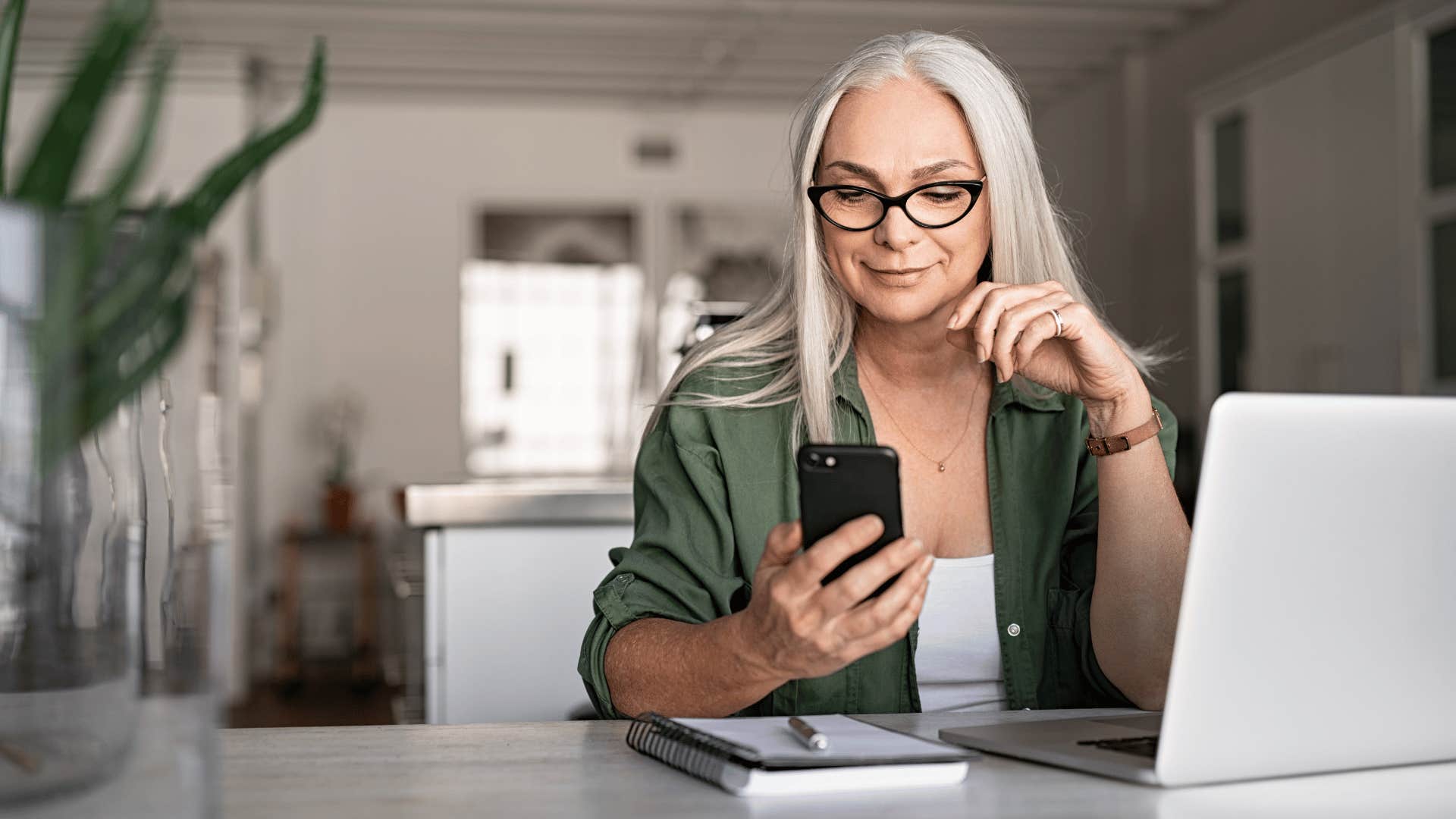 older woman looking at her phone while using her laptop