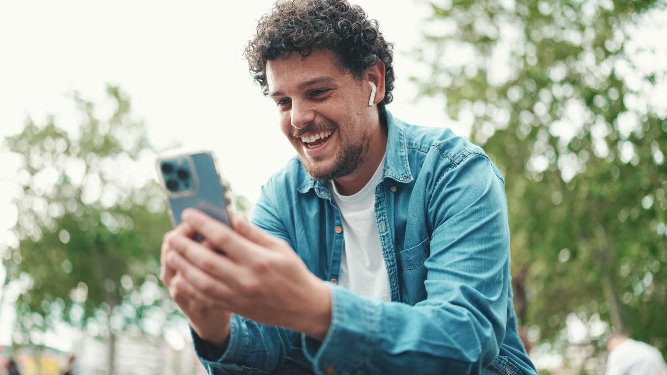 young bearded man in denim shirt sitting in wireless headphones making video call on mobile phone on busy street modern city background.