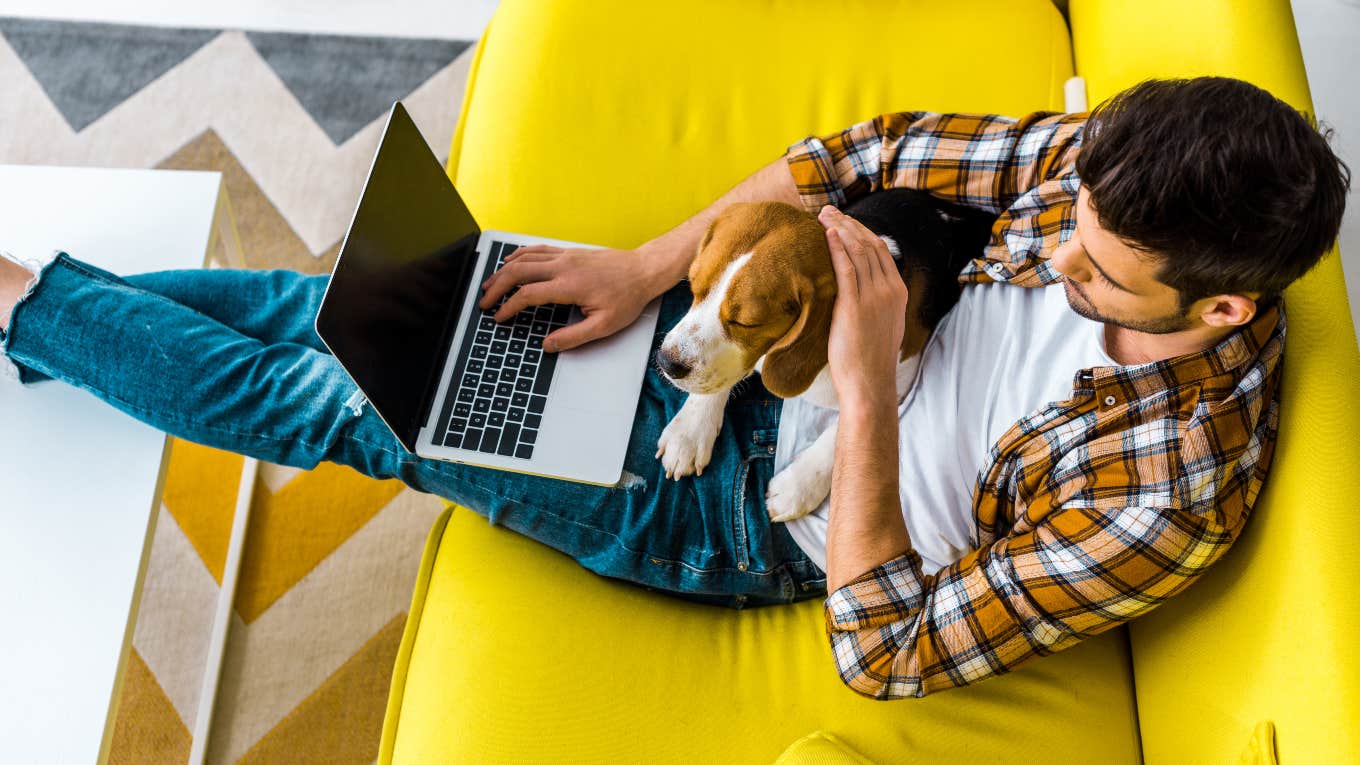 man sitting on couch working on laptop with his dog next to him