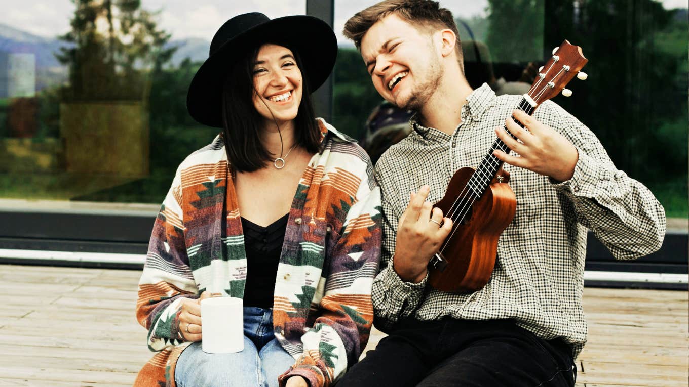 Husband and wife sitting outside laughing, husband playing guitar. 