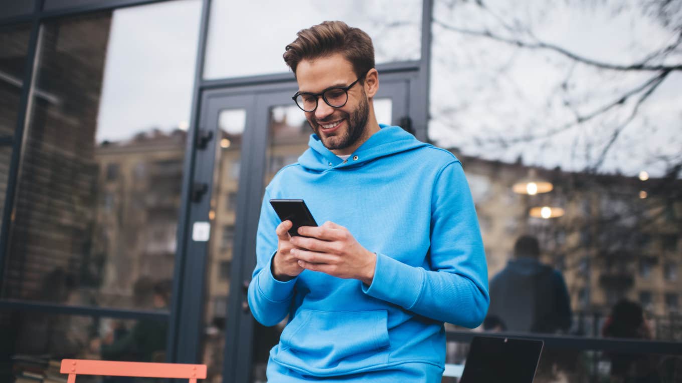 man wearing blue sweatshirt standing outside cafe texting