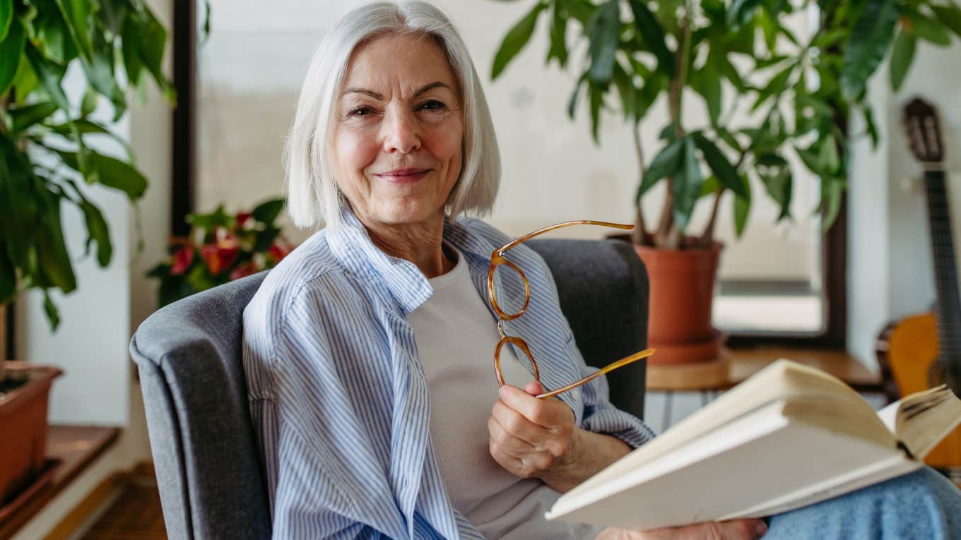 portrait of older woman holding glasses and reading book at home