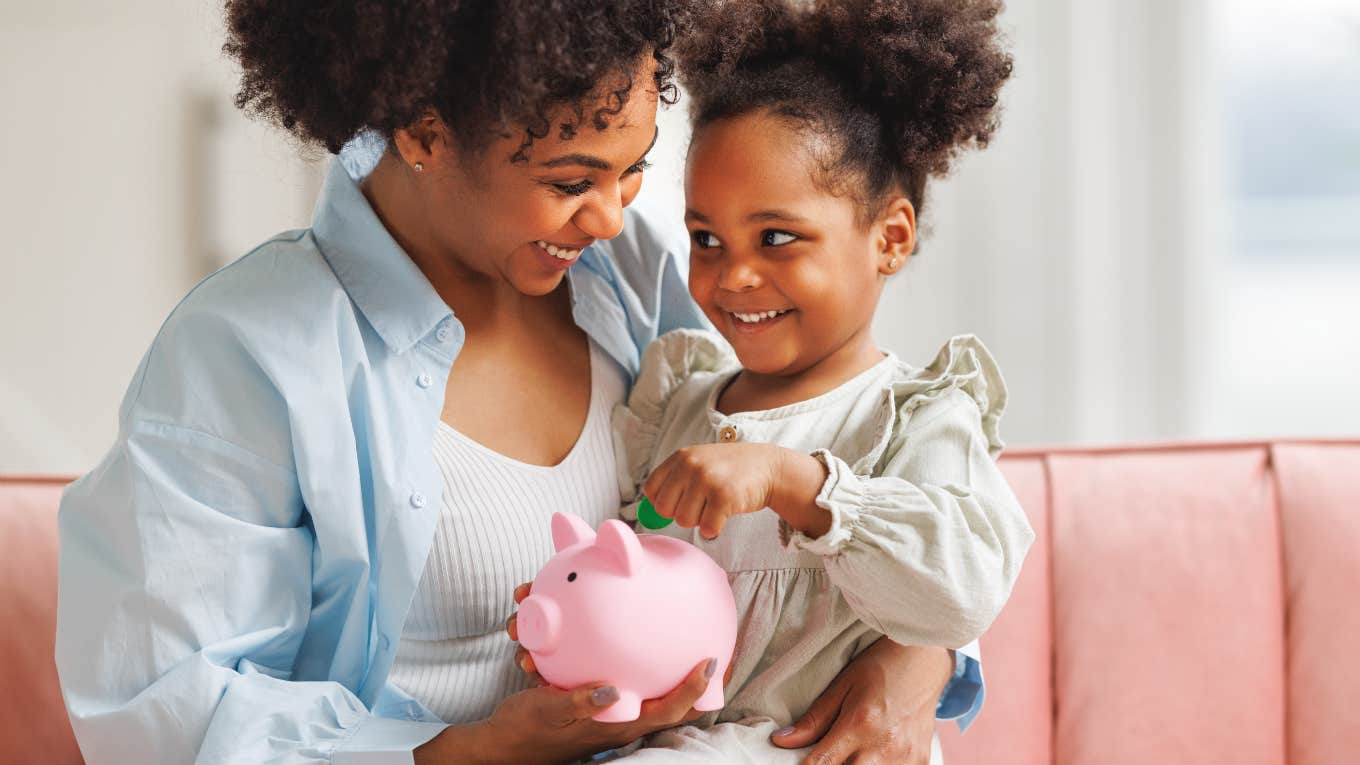 mom holding daughter in her lap putting money in piggy bank