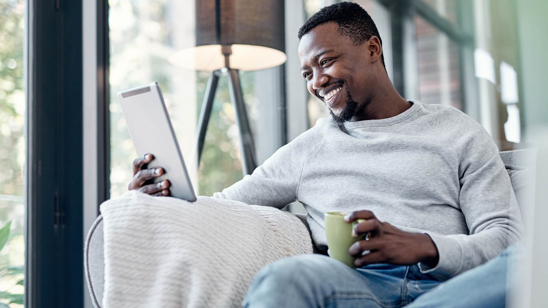 Man smiling while looking at a tablet and drinking coffee.