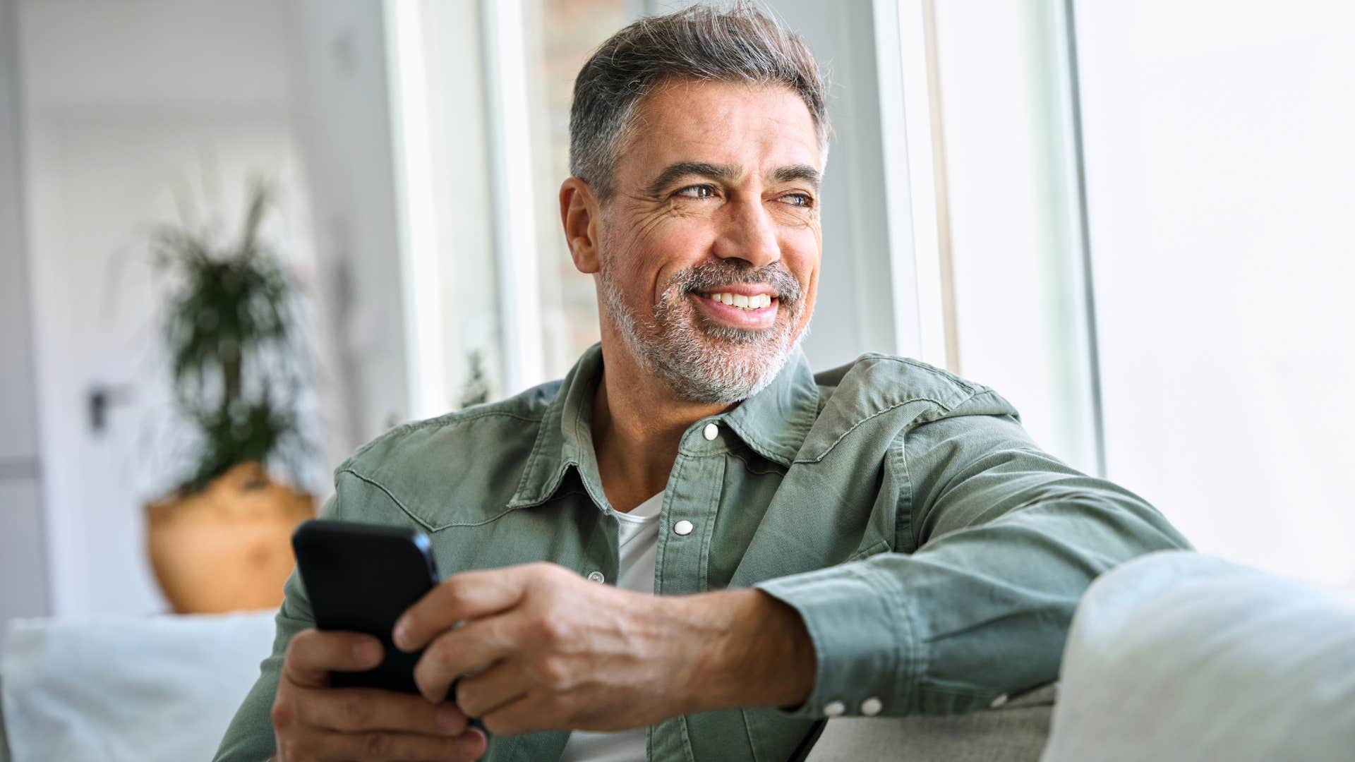 Older man smiling while texting and looking out his window.