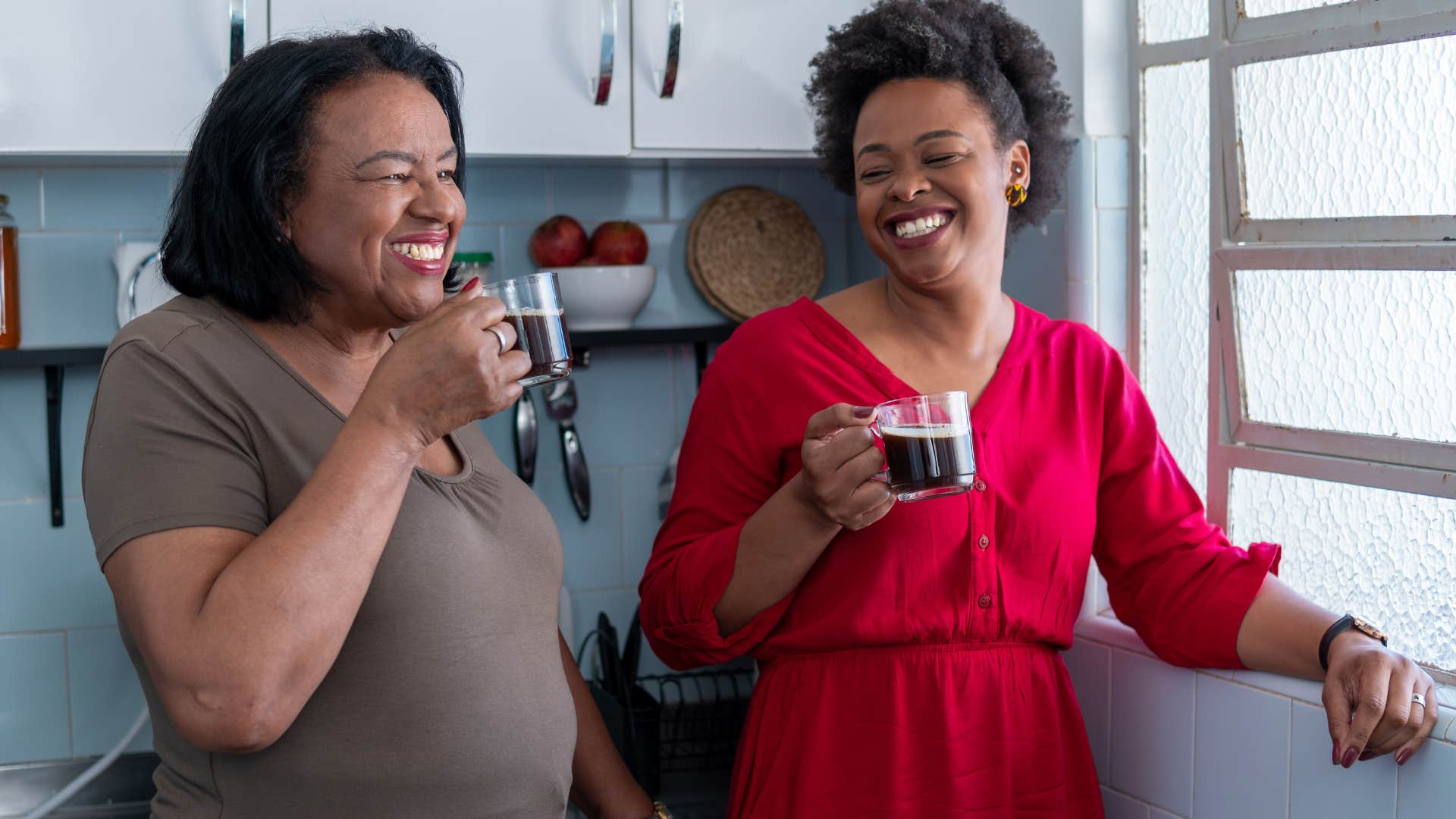 Adult daughter smiling and drinking coffee with her mother in the kitchen.