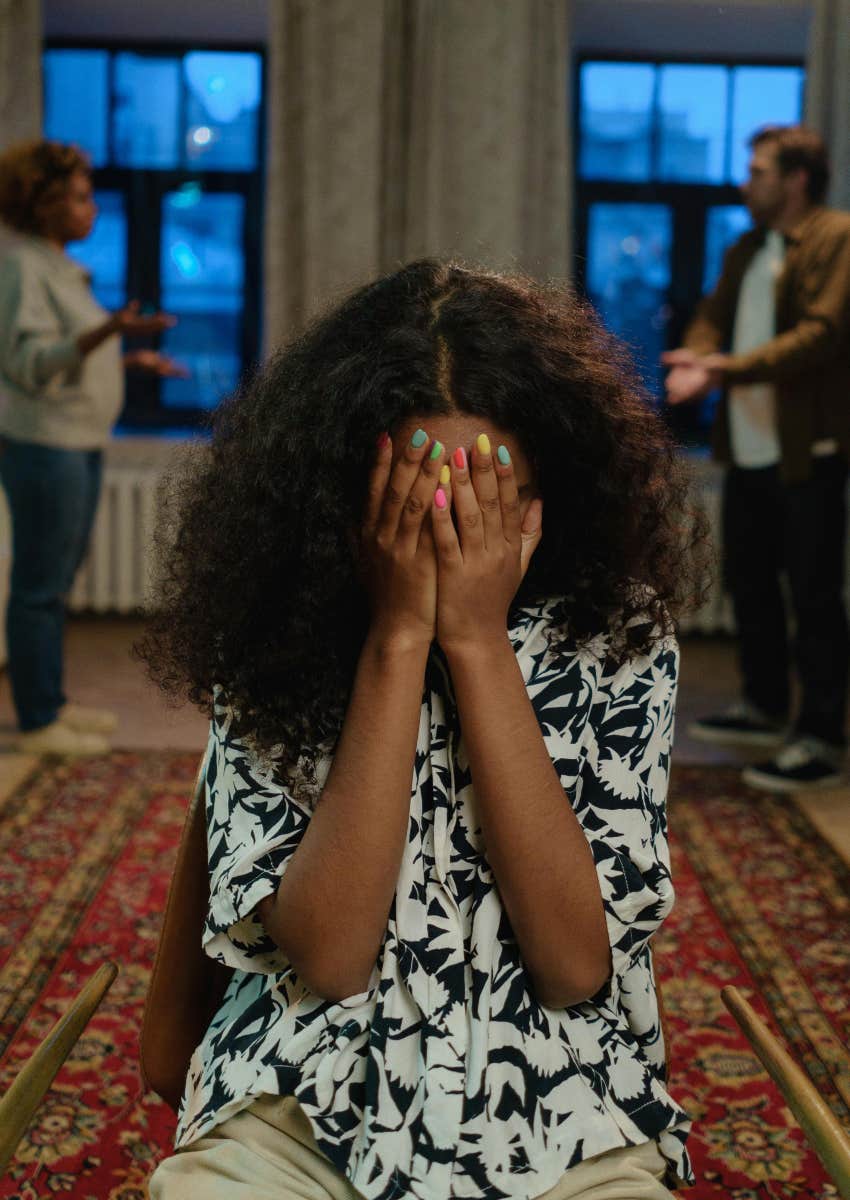 sad girl sitting on the ground while her parents fight in the background