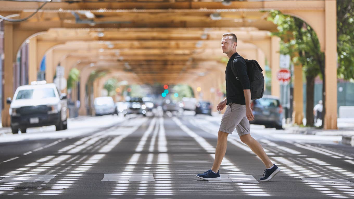 Side view of adult man with backpack walking on pedestrian crosswalk uder elevated railway of public transportation