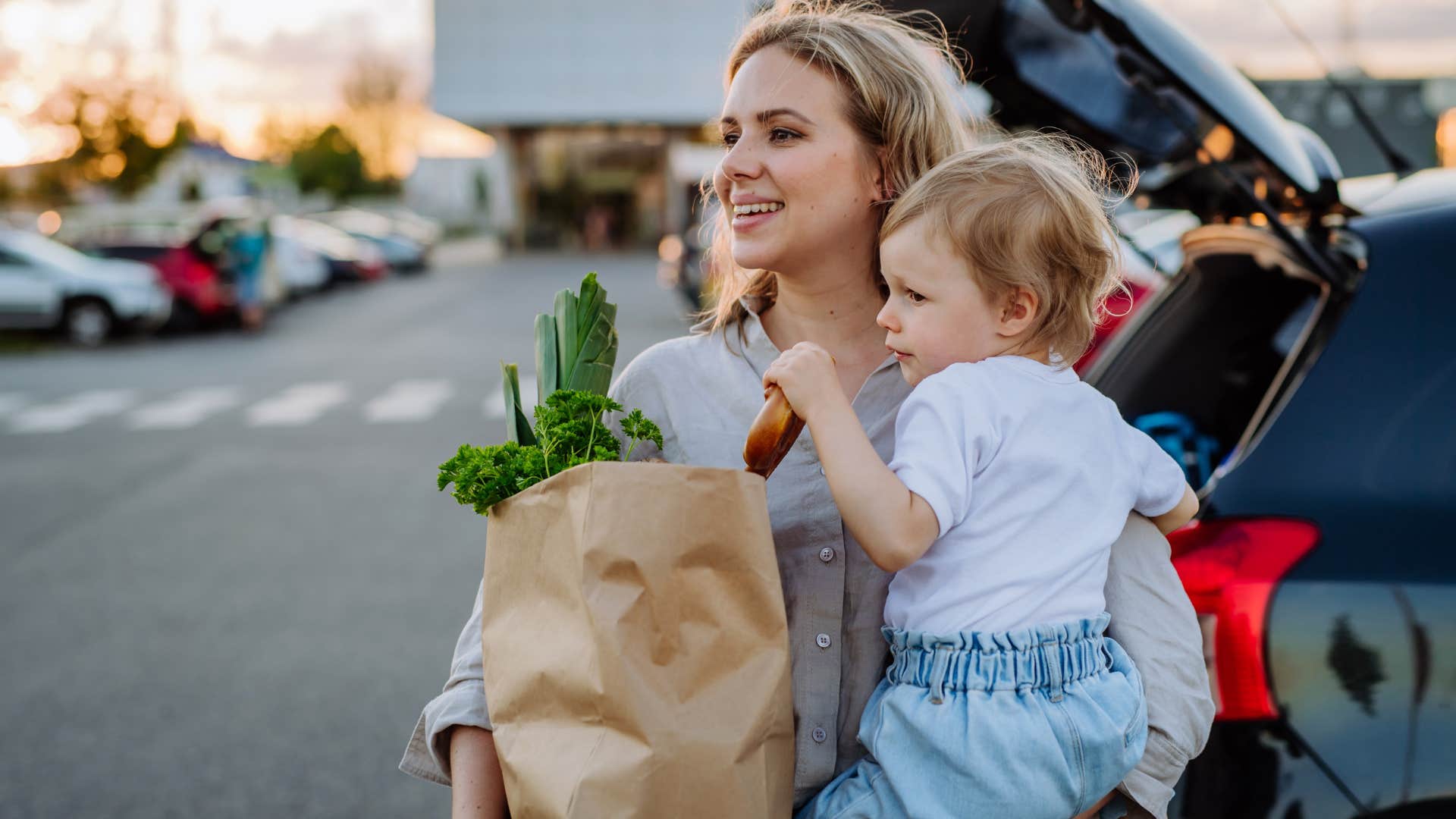 Woman smiling holding groceries and her baby in a parking lot. 