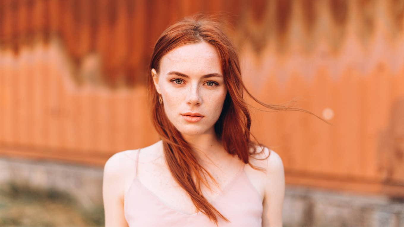 Close up portrait of young woman with long red hair and freckles posing outdoors on copper background. 